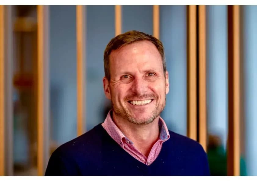 Portrait of Chris Blaine wearing a navy blue sweater with pink shirt collar in front of a blue, panelled wall